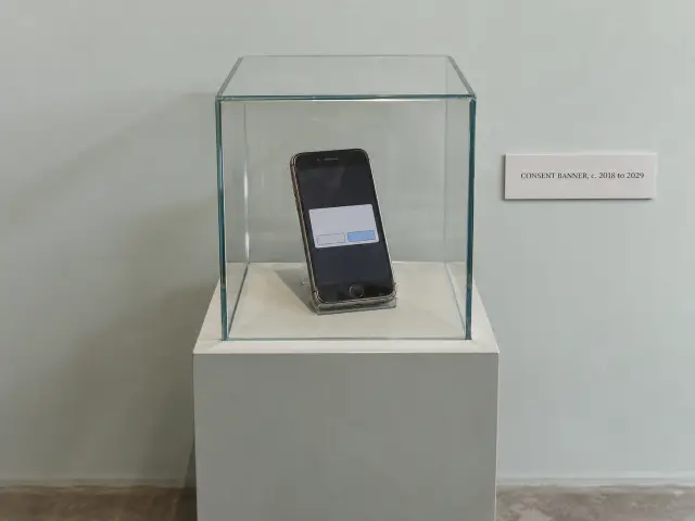 A museum display case holding an old phone with a pop-up box, beside a placard reading Consent banner, c. 2018 to 2029.